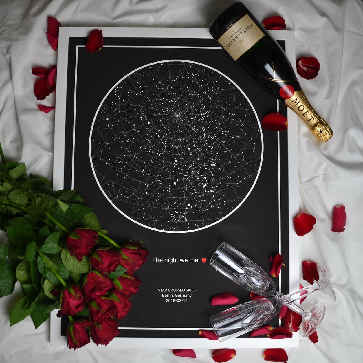 Black star map in a white frame lying on a bed with red roses, champagne and two glasses